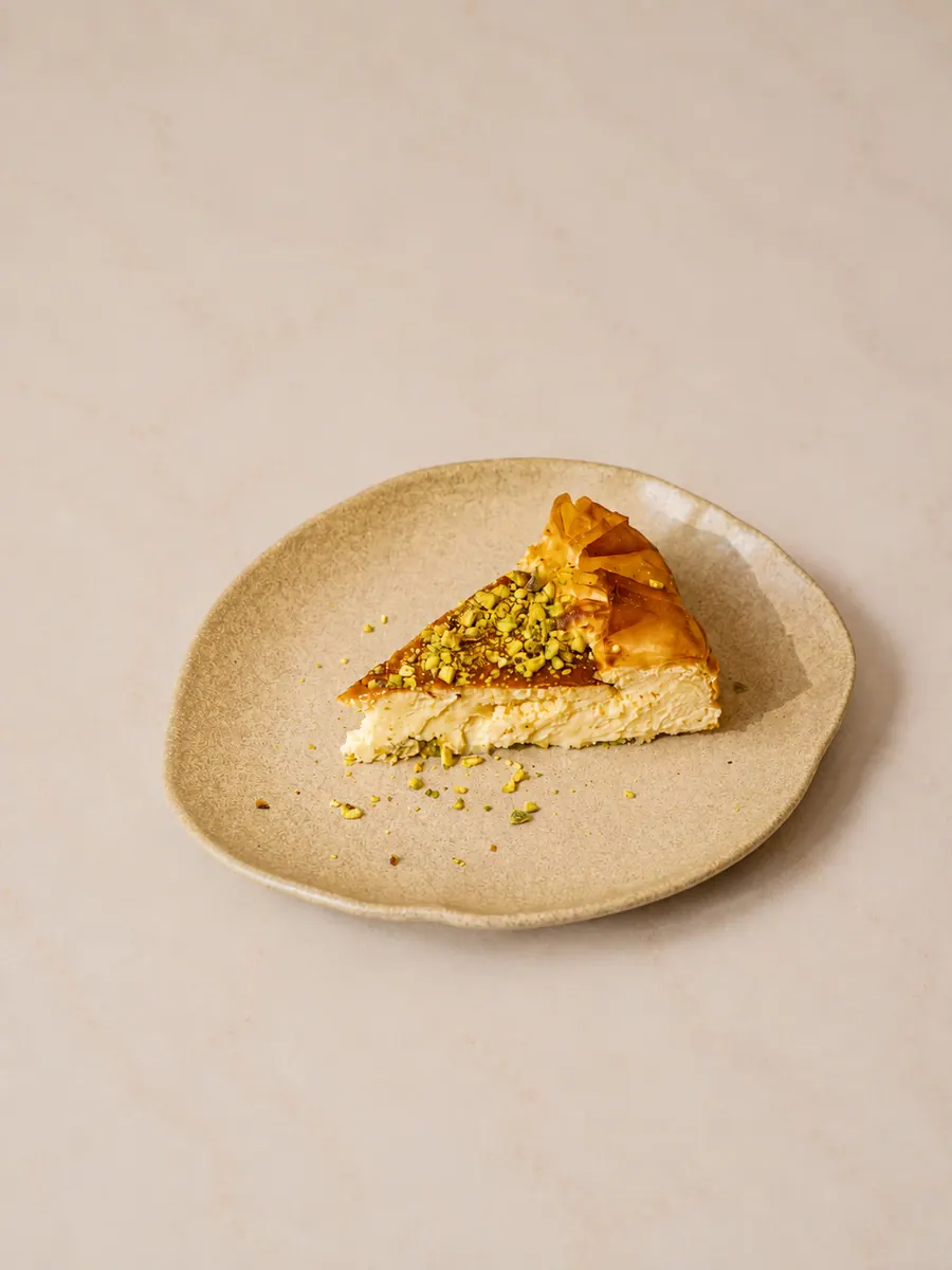 Al Reem baklava cheesecake: a wedge of baked cheesecake on a speckled beige plate, its top glazed golden under a layer of filo pastry and heaped with crushed pistachio.