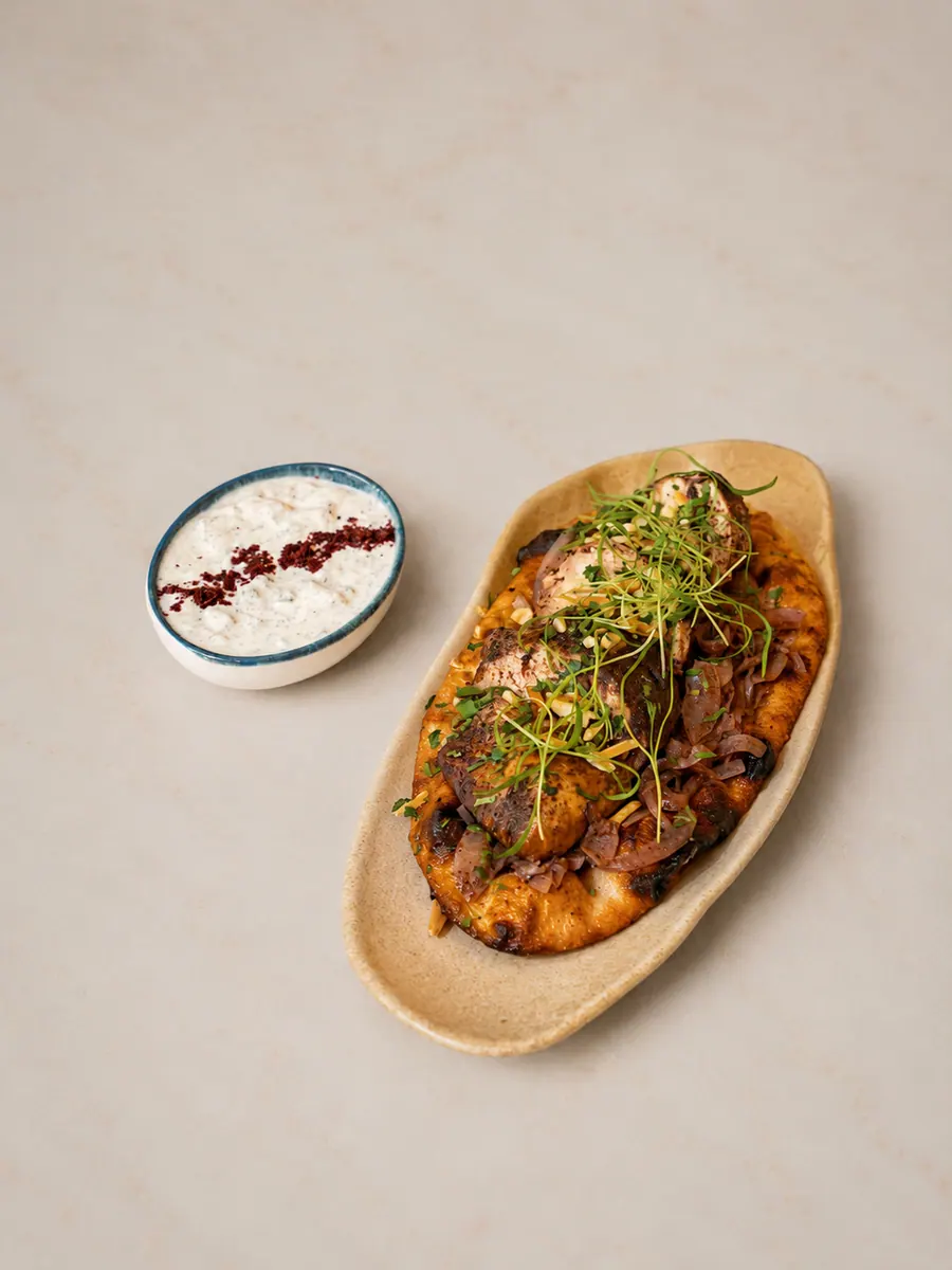 Al Reem traditional musakhan: chicken and dark caramelised onion piled on an oval of taboon flatbread, topped with green shoots and chopped parsley, with a small bowl of yogurt dusted with sumac beside it.