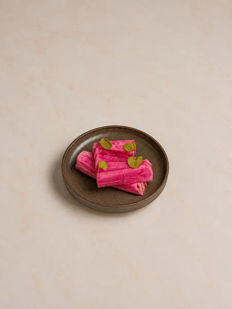 Al Reem pickles: bright pink batons of pickled turnip, coloured by beetroot, stacked on a small dark brown ceramic plate with a few small green leaves.