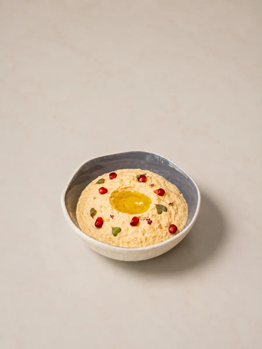 Al Reem moutabbal: a shallow bowl of pale, creamy charcoal-roasted eggplant dip with a pool of olive oil in the centre, scattered with pomegranate seeds and small green leaves.